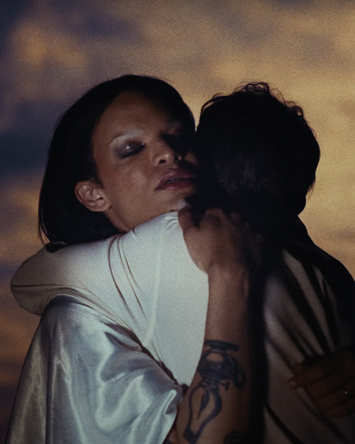 Still frame of two figures embracing one another against as gradient sunset background